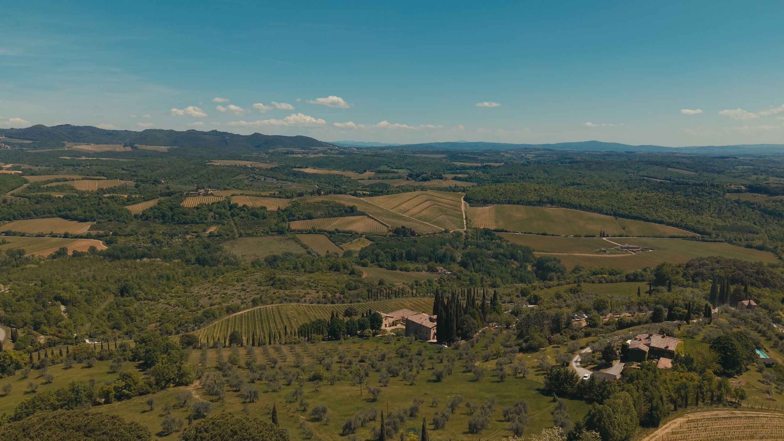 Aerial view of Tuscan countryside with rolling hills, vineyards, farmhouses, and a wind turbine under blue sky.