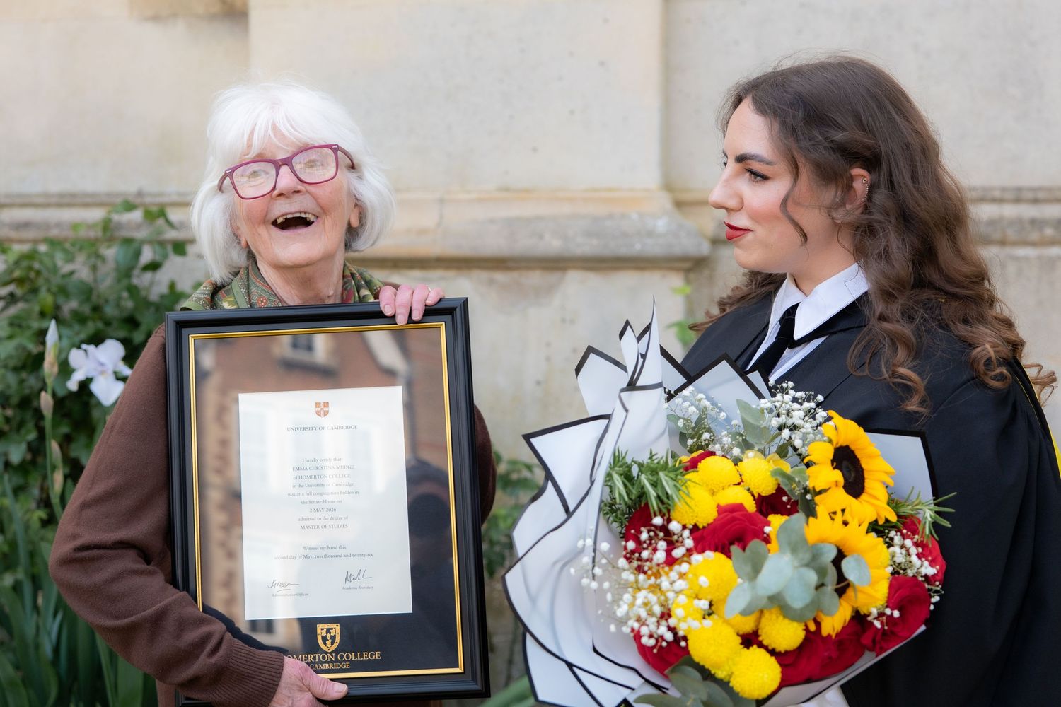 Elderly woman laughing holding framed certificate beside young graduate in black gown with colorful flower bouquet.