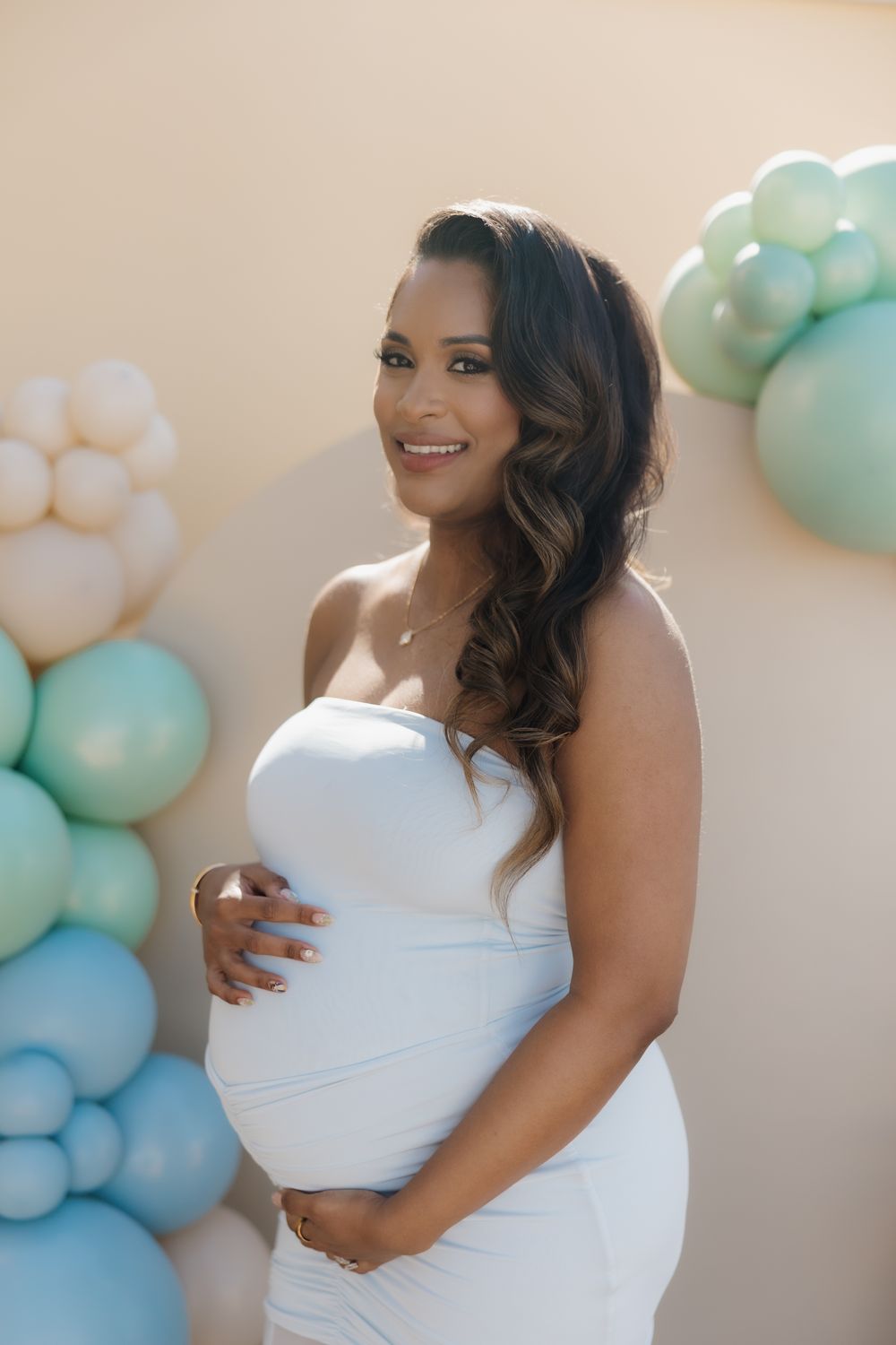 Smiling pregnant woman in white strapless dress holding baby bump at baby shower with blue and green balloon backdrop.