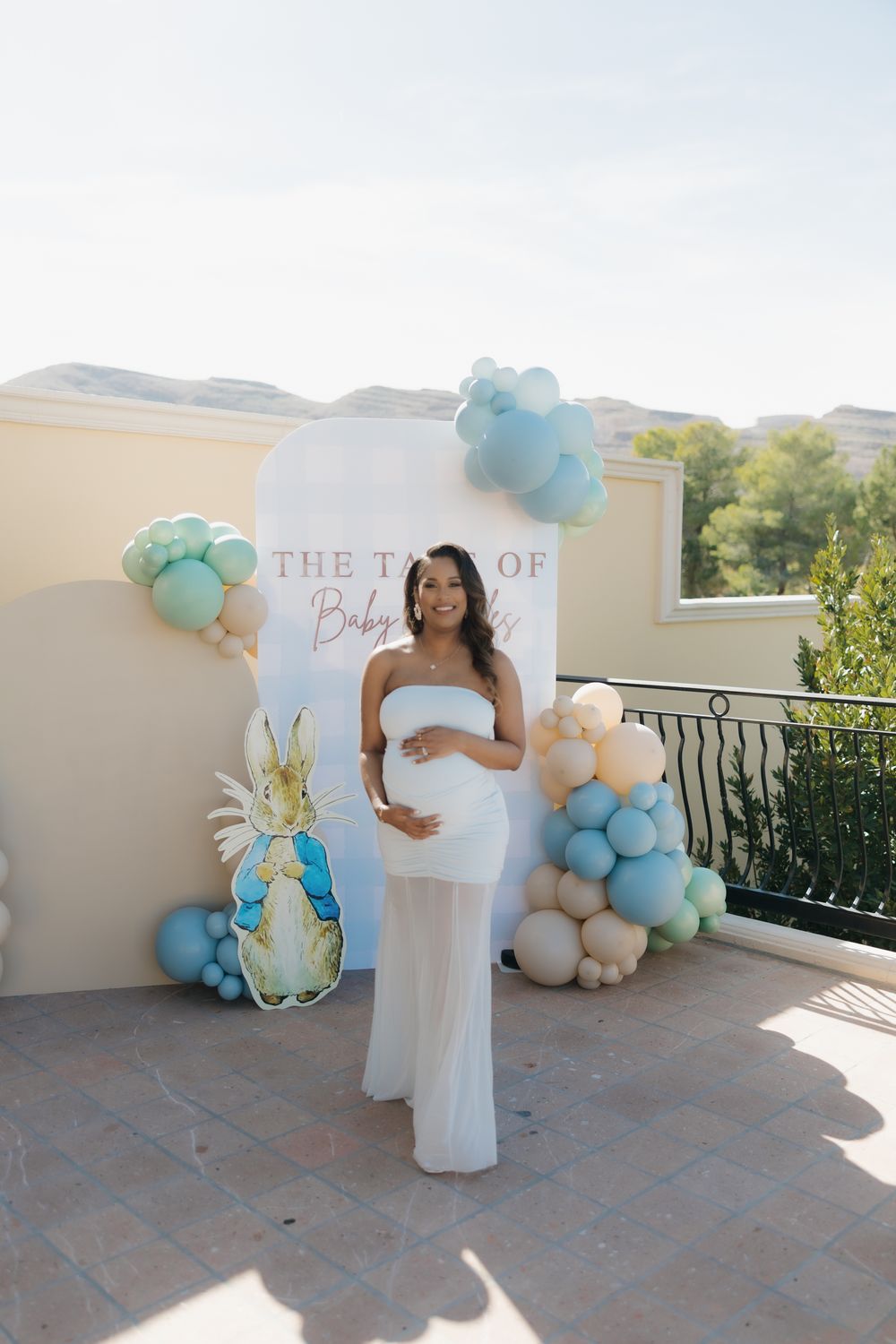Pregnant woman in white dress poses at Peter Rabbit baby shower backdrop with pastel balloons.