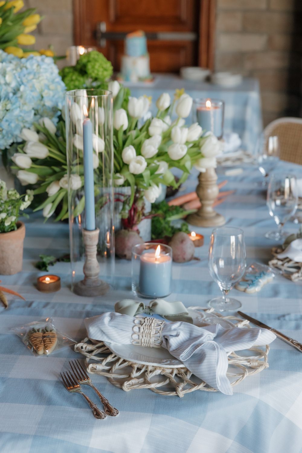 Elegant blue gingham Easter table setting with white tulips, hydrangeas, candles, and decorative place settings.
