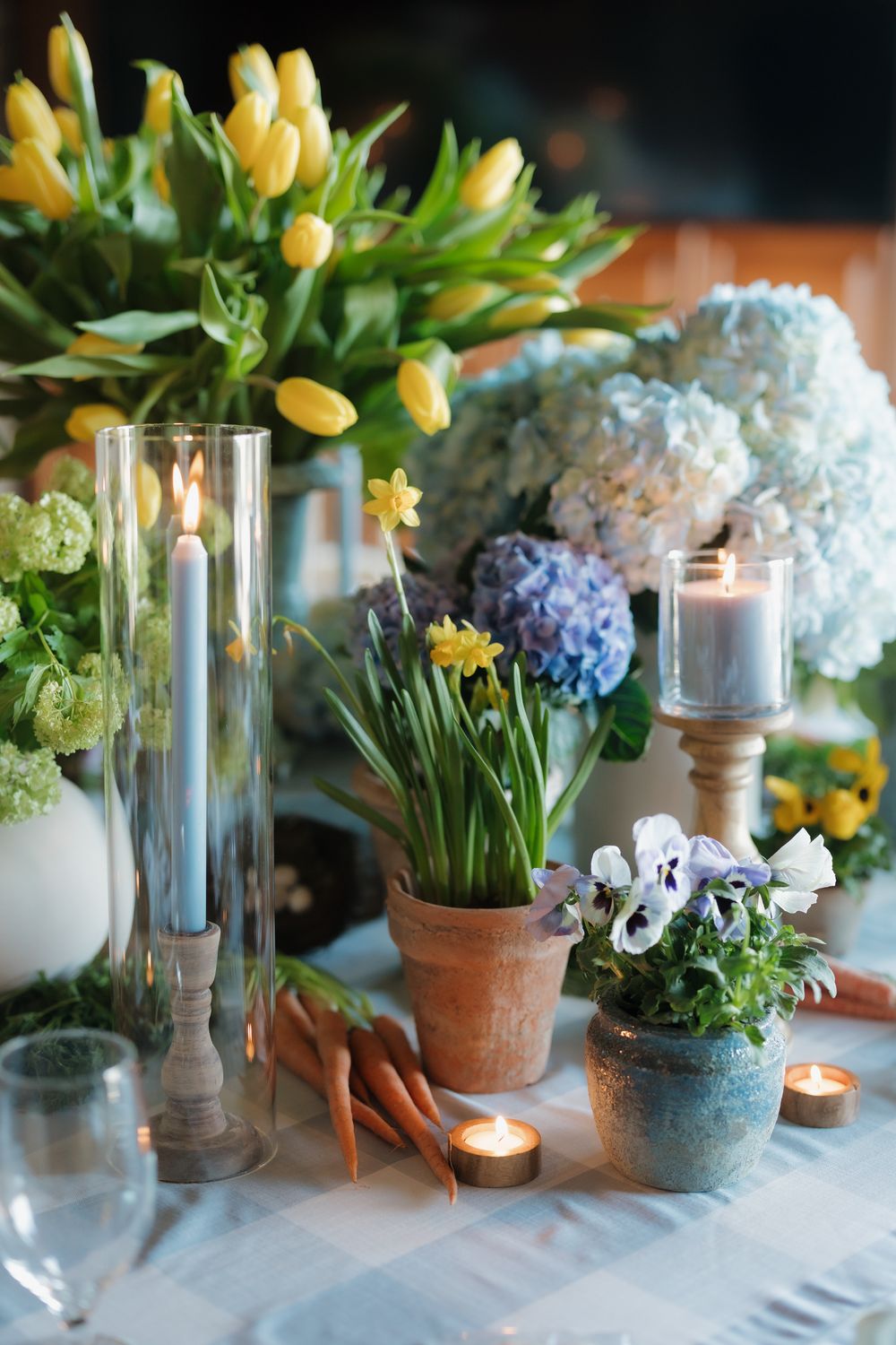 Spring tablescape featuring yellow tulips, blue hydrangeas, potted pansies, carrots, and lit blue candles.