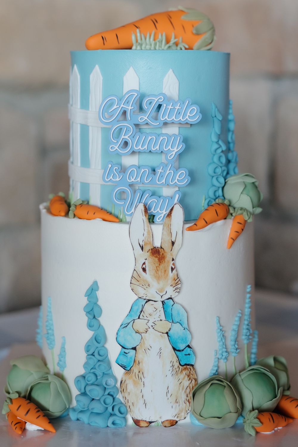 Two-tier Peter Rabbit baby shower cake with carrots, cabbages, and “A Little Bunny is on the Way” text.