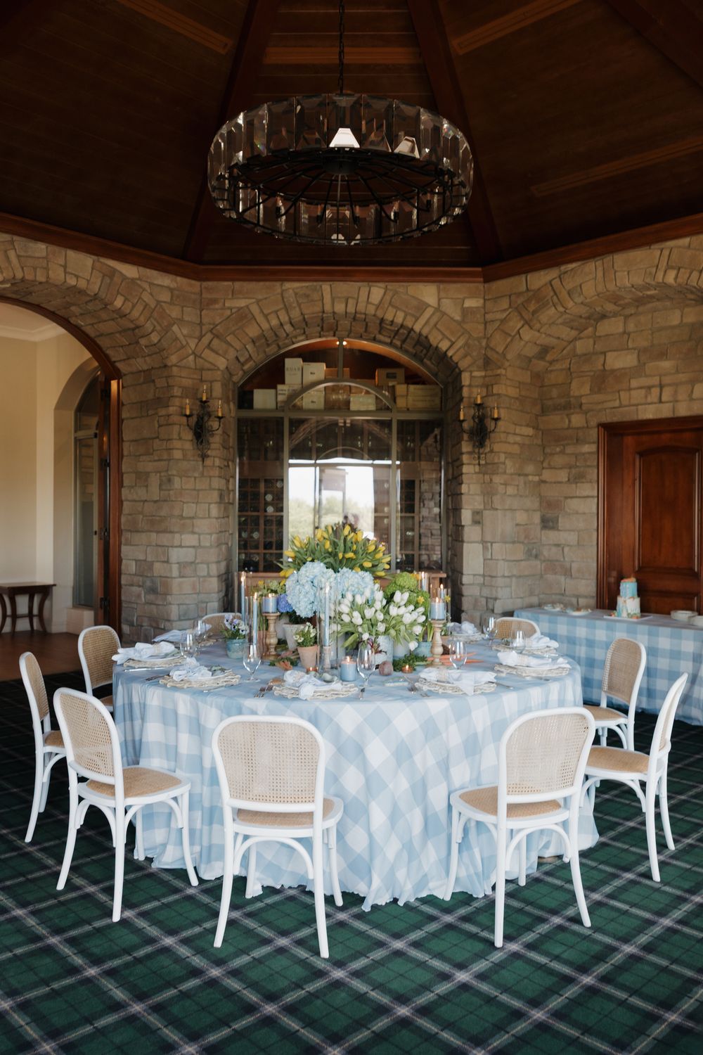 Elegant round dining table with blue plaid tablecloth, white chairs, floral centerpiece in stone-walled event venue.