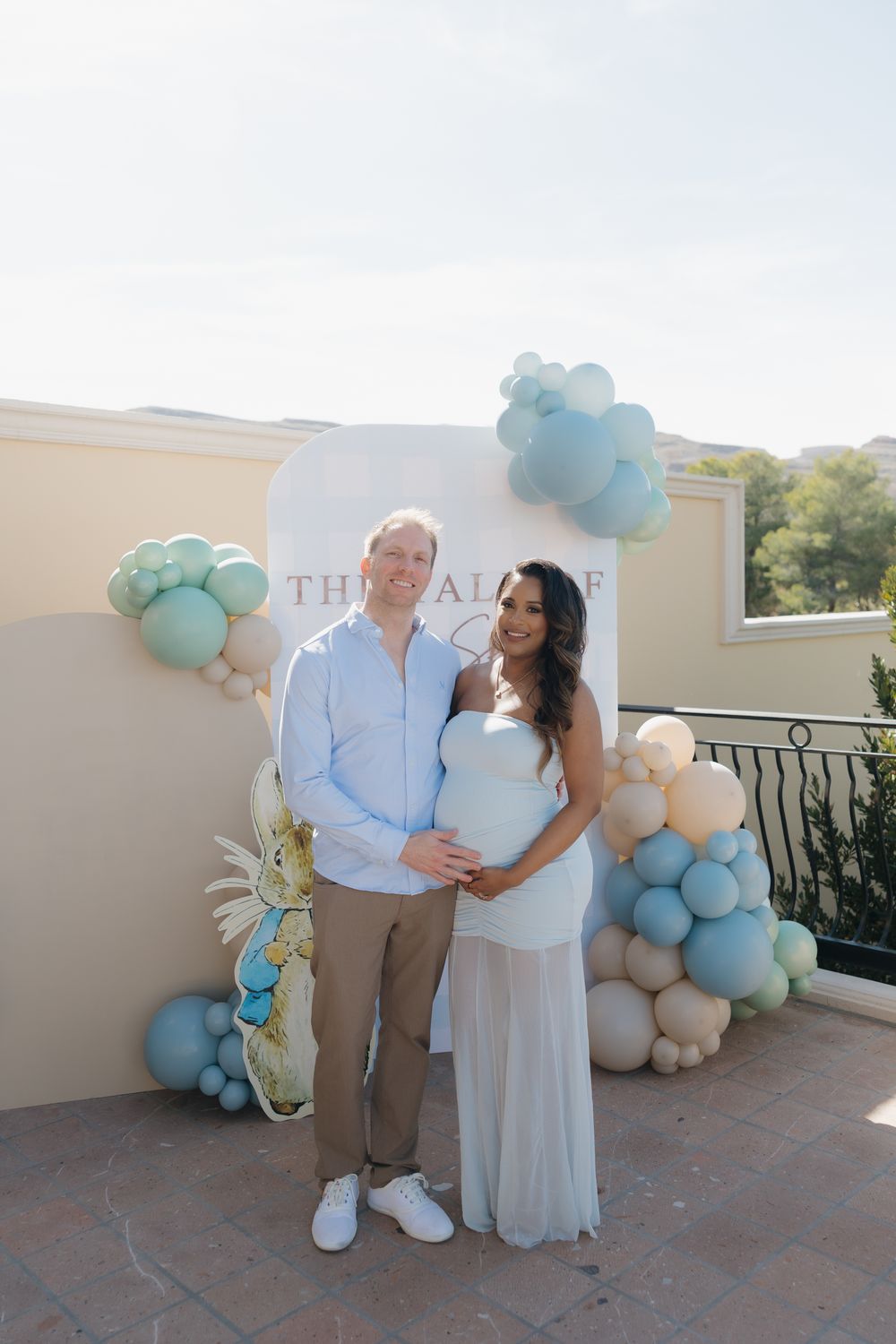 Couple poses at baby shower with blue and white balloon decorations outdoors.