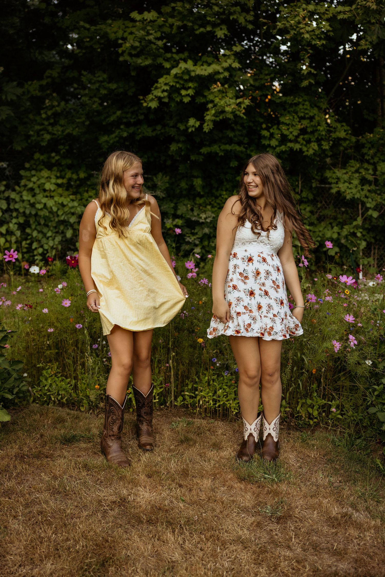 Two young women in summer dresses and cowboy boots smile at each other in a wildflower garden.