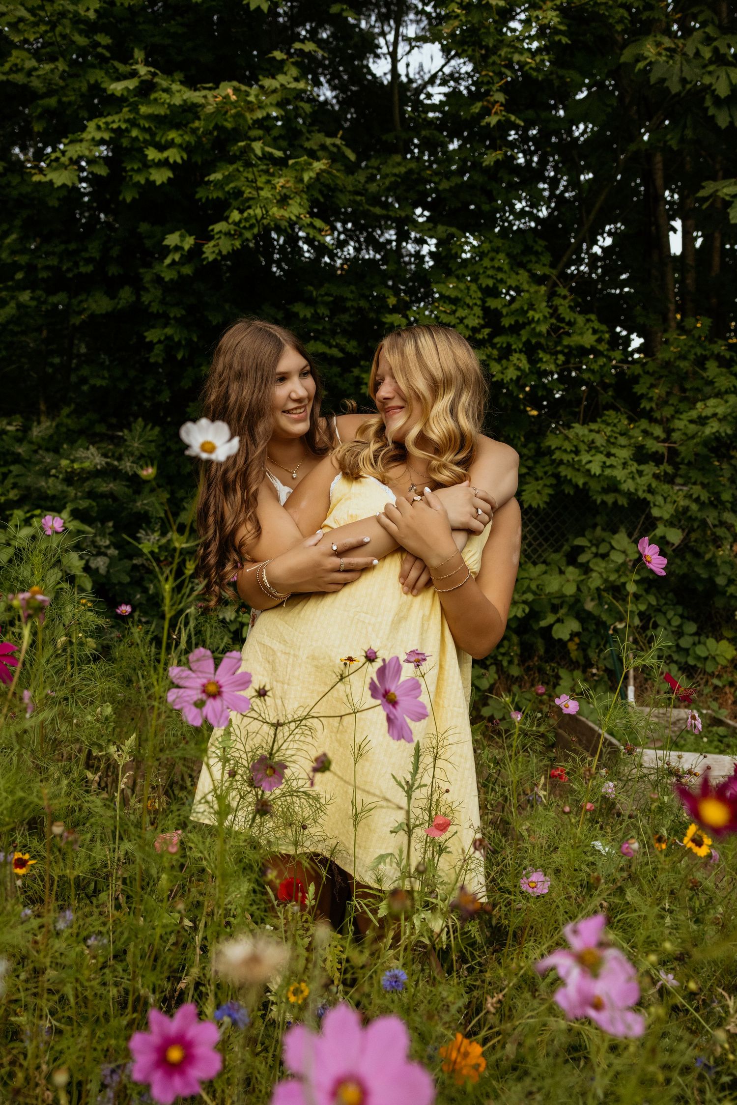 Two young women in yellow dresses embrace and laugh together in a colorful wildflower garden with lush green trees.