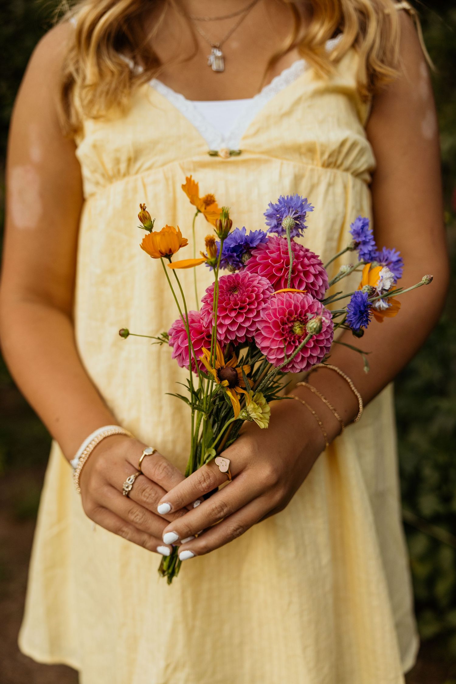 Woman in yellow dress holding a colorful wildflower bouquet with dahlias, cornflowers, and marigolds, wearing gold rings.