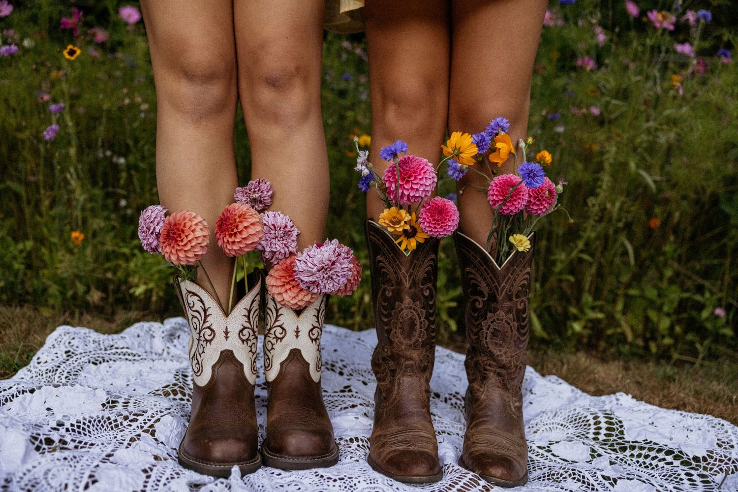 Two pairs of legs in cowboy boots filled with colorful dahlias and wildflowers on a lace blanket in a garden.