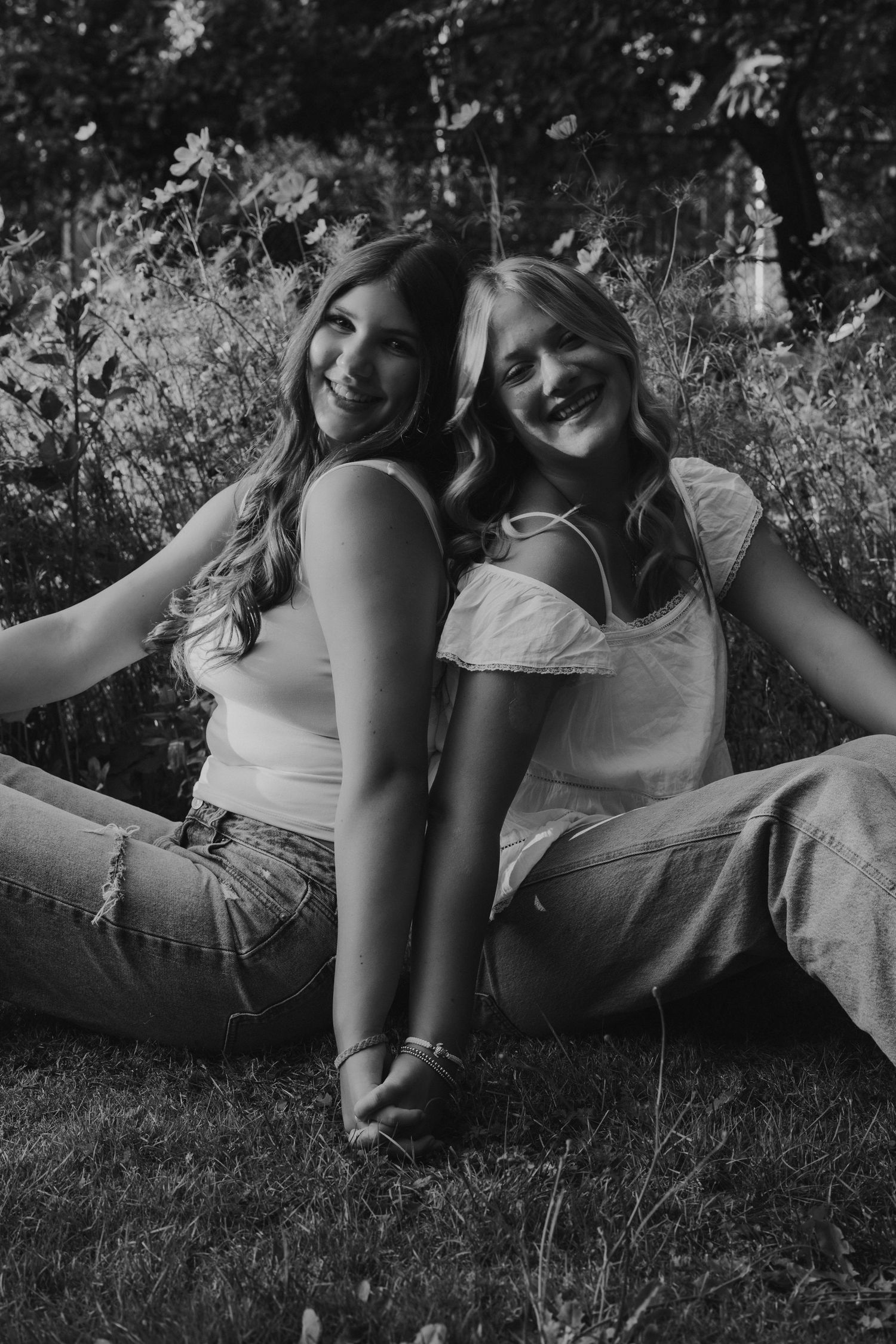 Two smiling young women sitting back-to-back on grass in a black and white outdoor portrait photo.
