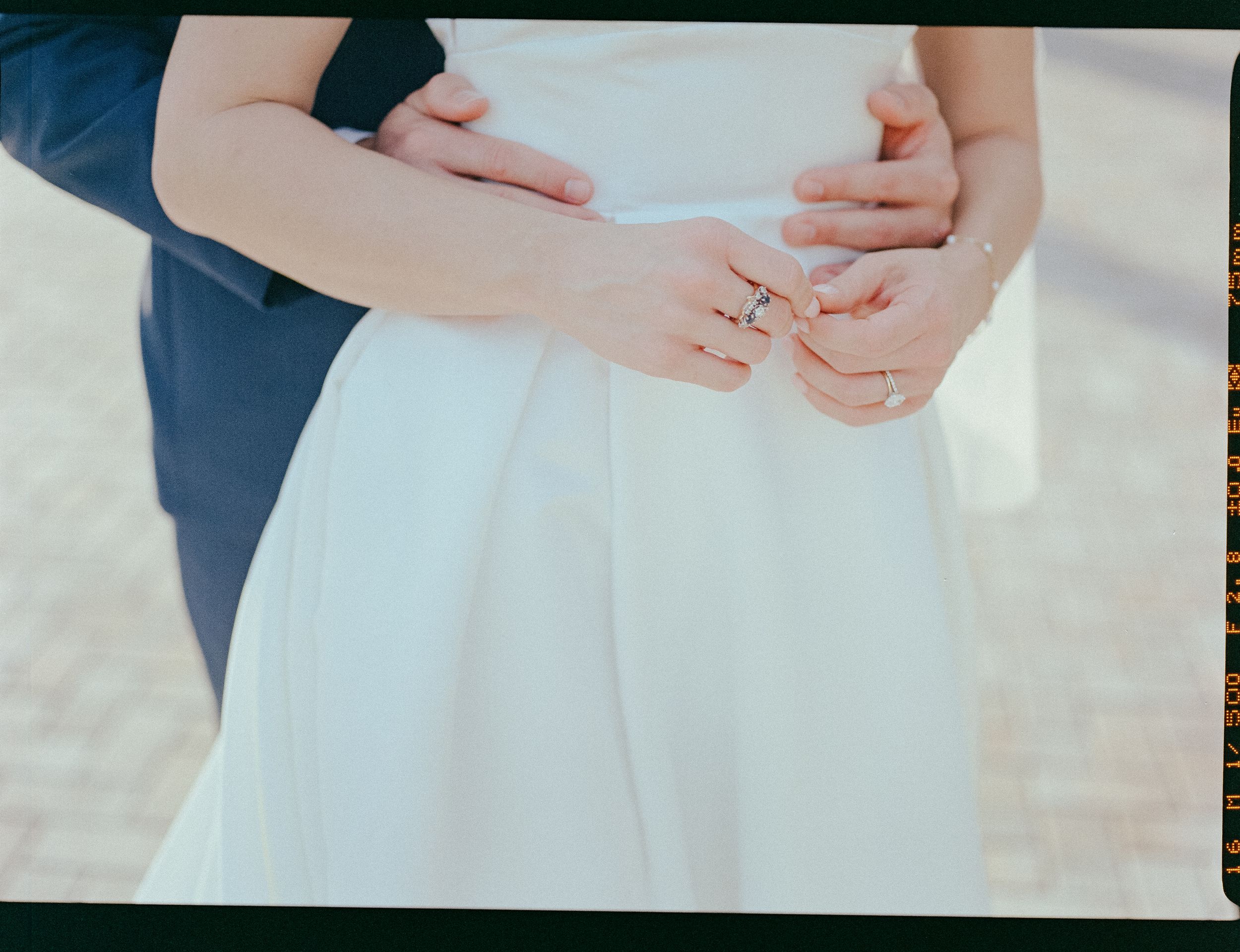 Bride and groom holding hands showing wedding rings, bride in white dress, groom in navy suit, outdoor wedding photo.