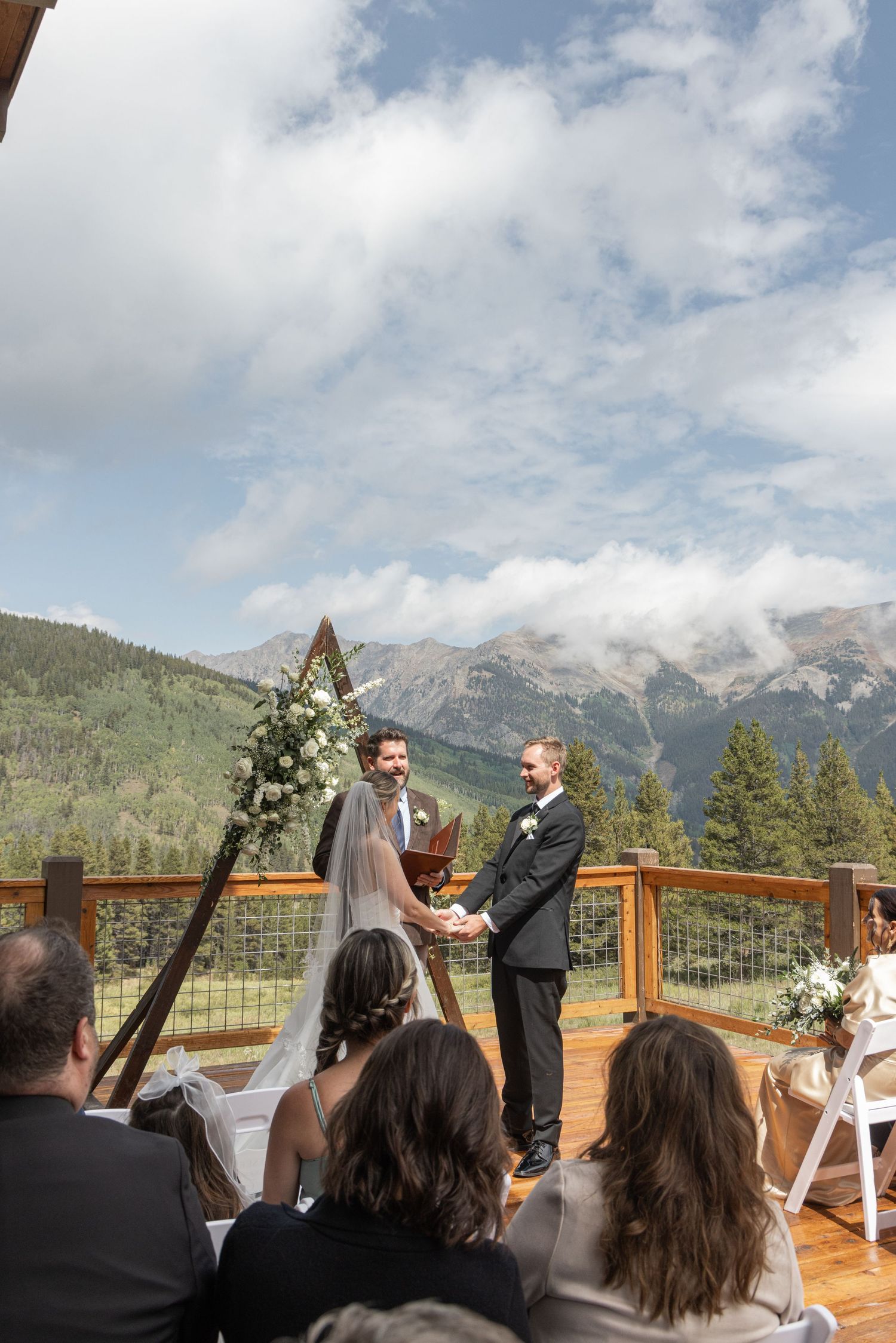 Outdoor mountain wedding ceremony on a wooden deck with couple exchanging vows before stunning Rocky Mountain backdrop.
