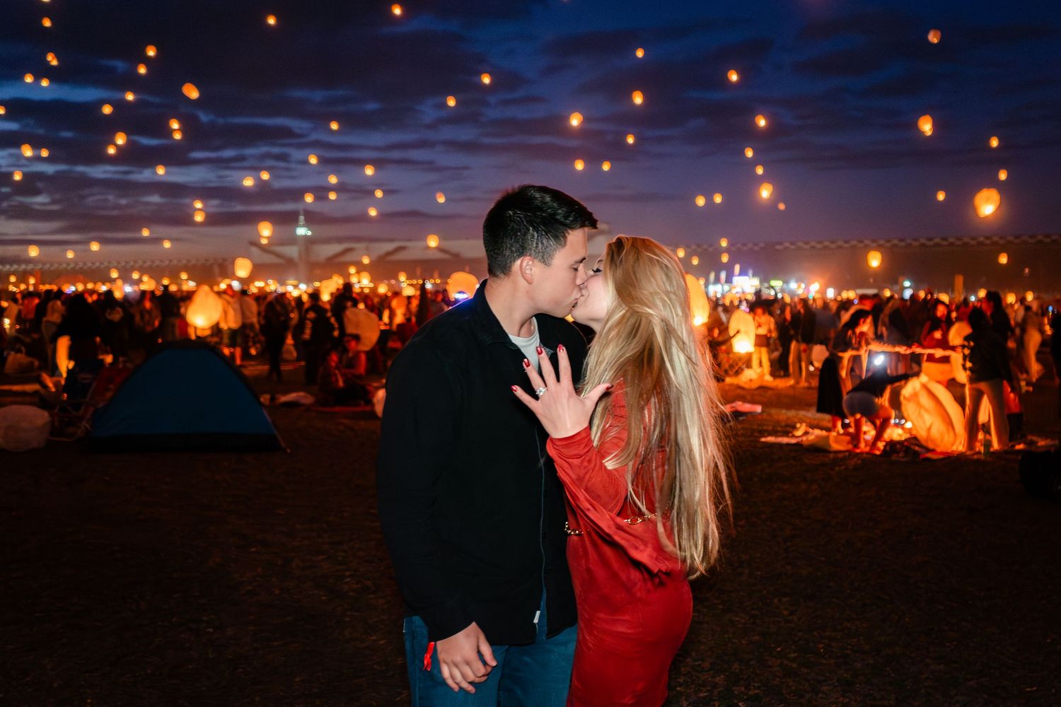 Couple kissing at a romantic night festival with glowing sky lanterns floating above a crowd under a twilight sky.