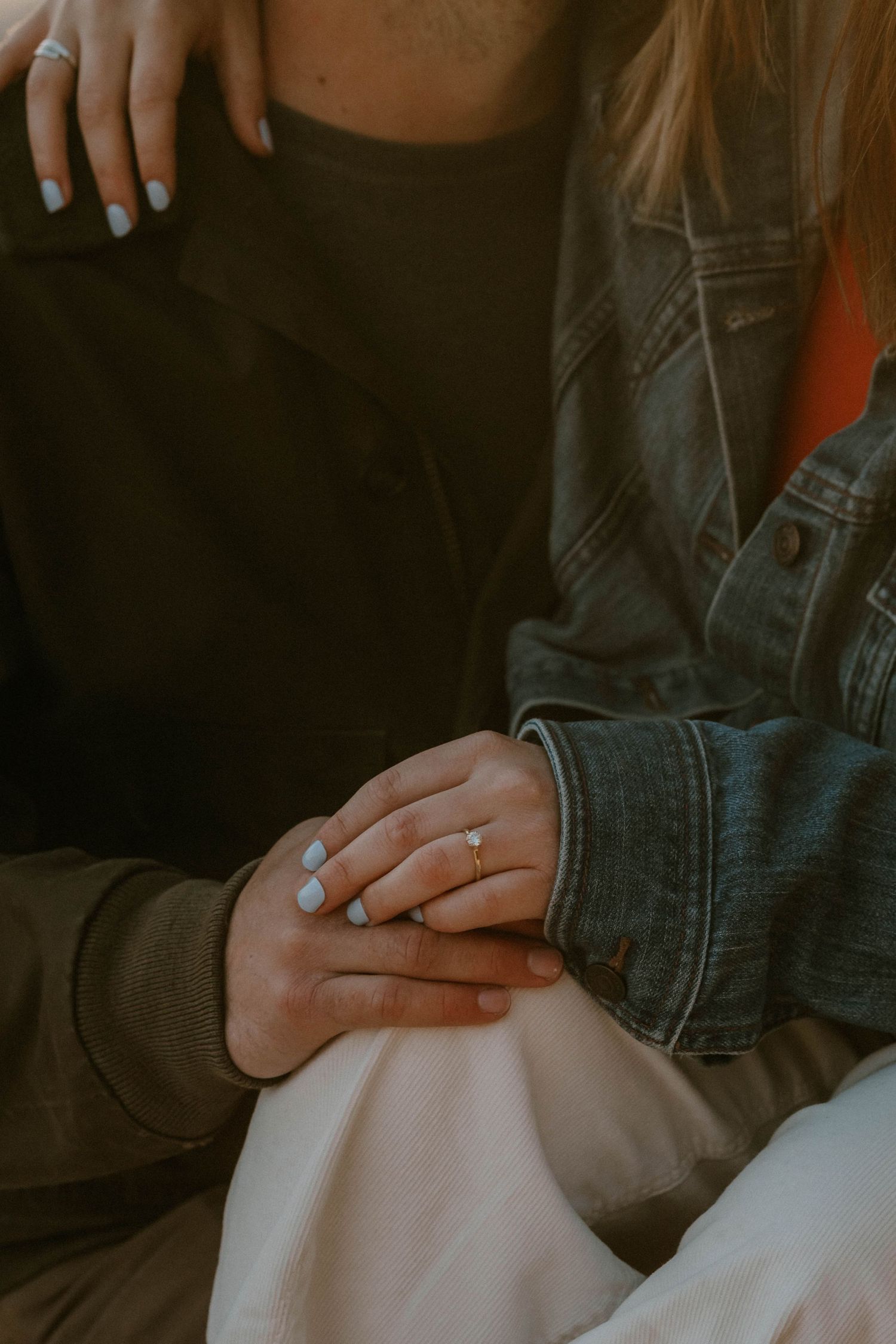 Couple holding hands with engagement ring visible, wearing denim jacket and black outfit, romantic close-up photo.