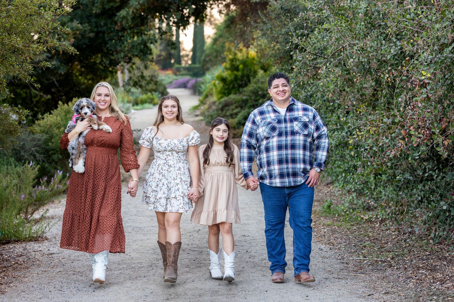 Family of four walking on a garden path, smiling, with a dog, dressed in casual western attire.