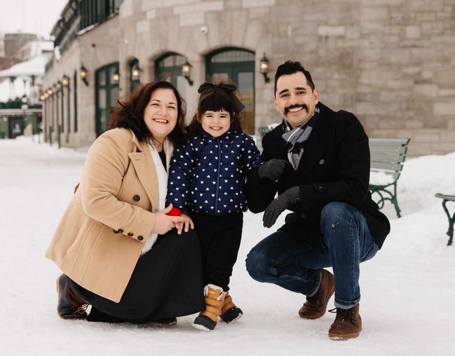 Family of three posing together in the snow outside a historic building in winter, smiling warmly at the camera.