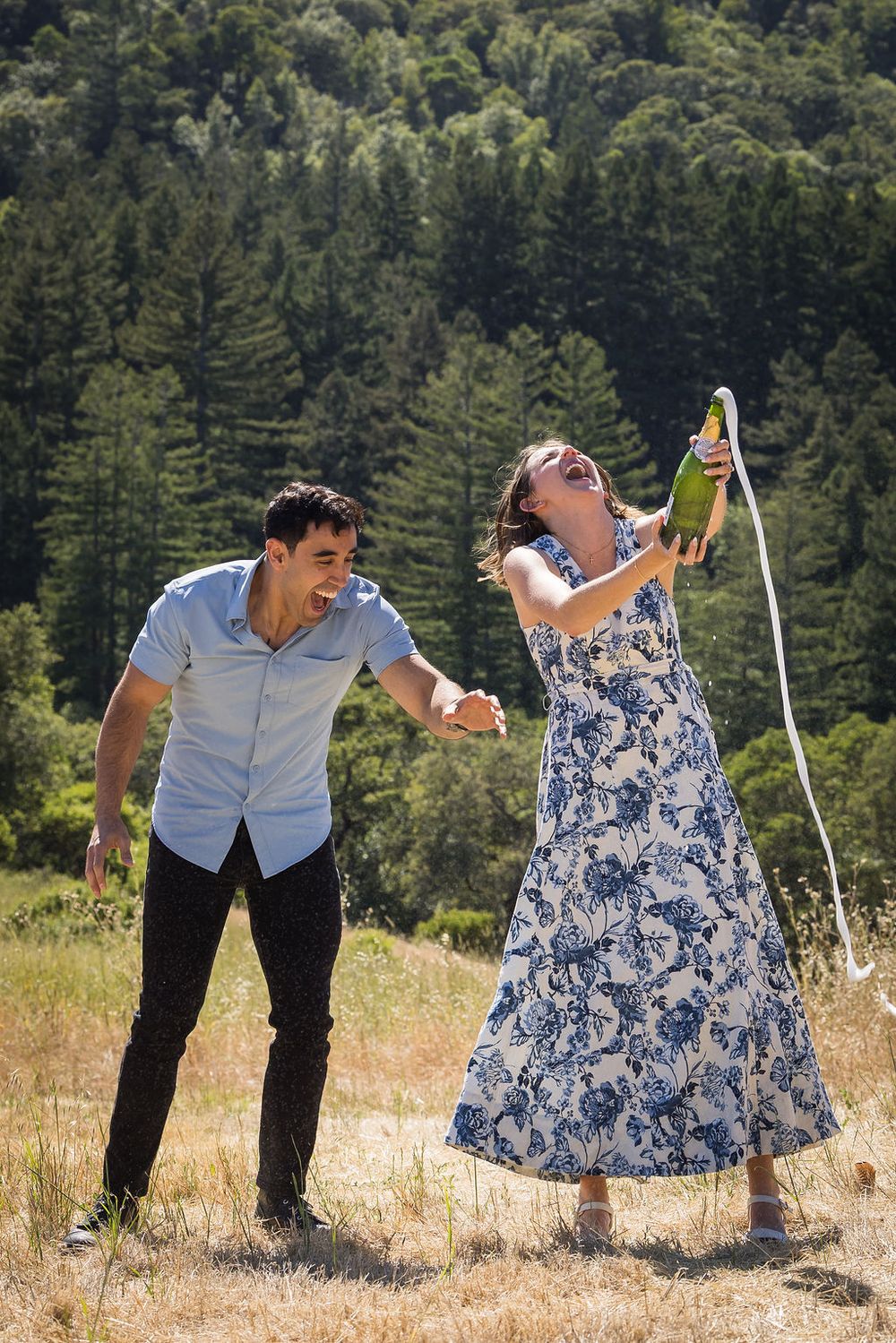Joyful couple celebrating outdoors as woman pours champagne bottle in sunny green hillside setting.