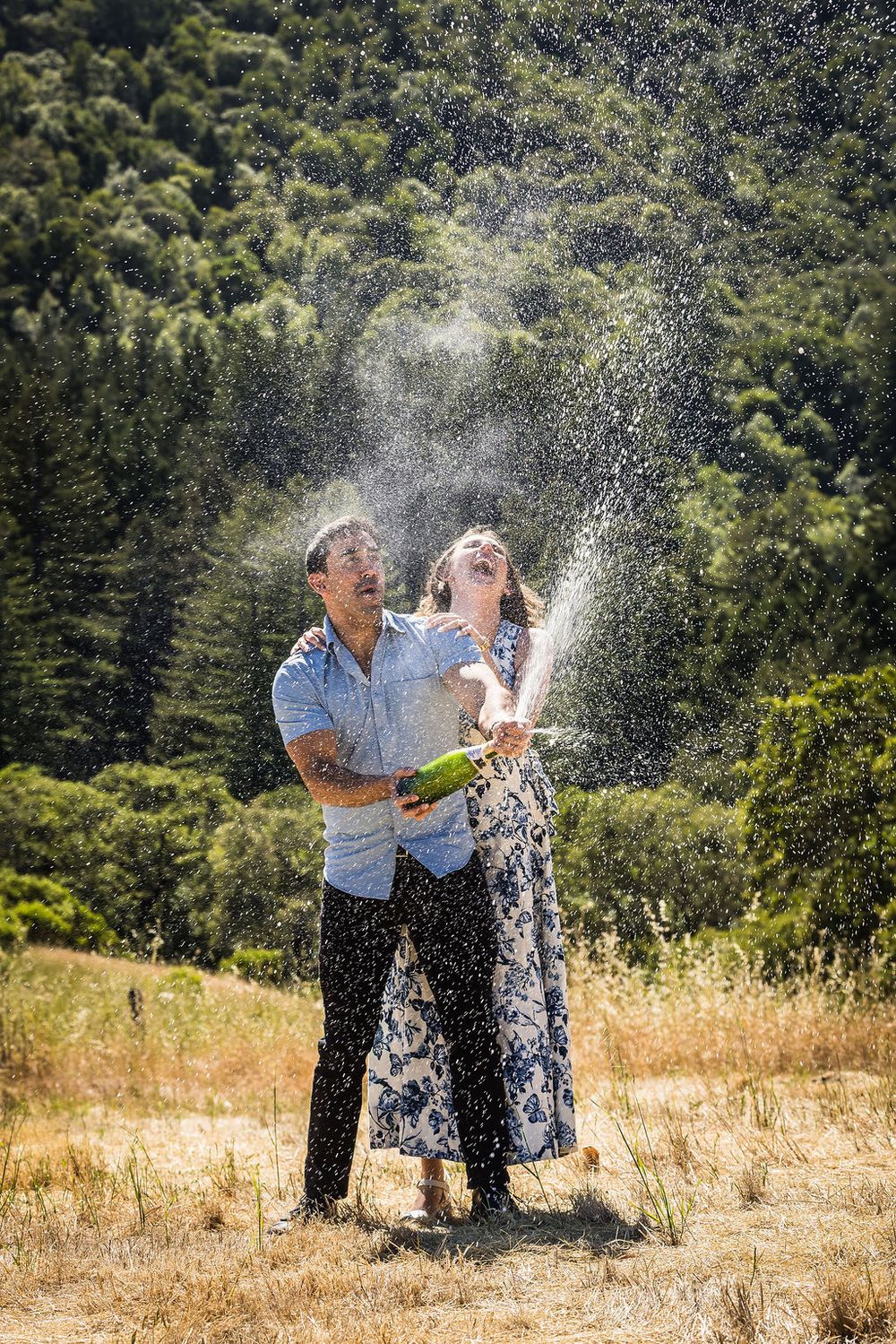 Joyful couple popping champagne bottle outdoors with green trees in background on sunny day.