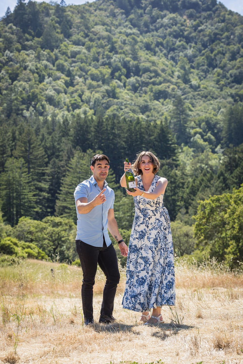 Couple celebrating outdoors with champagne bottle on sunny hillside with lush green forested mountain backdrop.