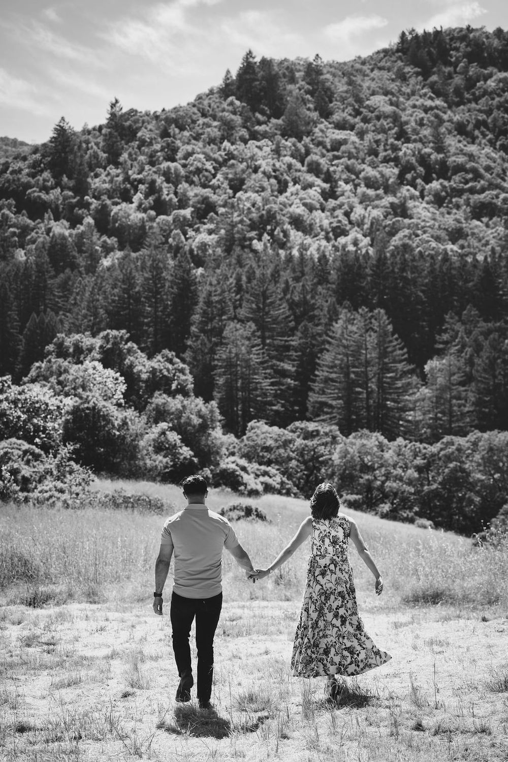 Black and white photo of a couple holding hands in a meadow with a lush forested hillside behind them.