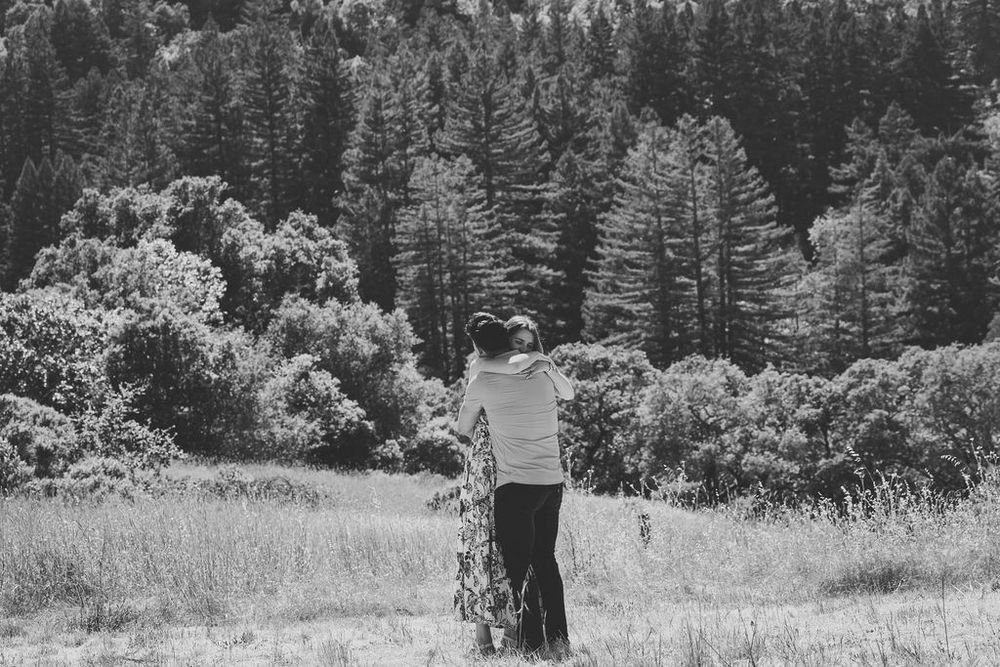 Black and white photo of a couple embracing in an open field surrounded by tall pine trees.