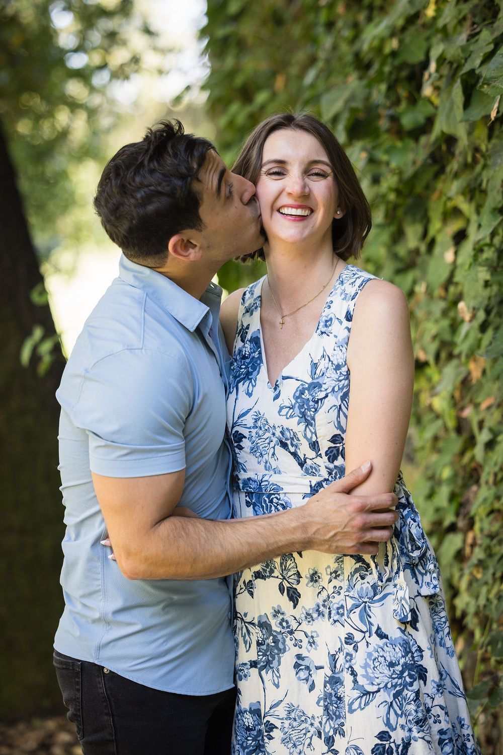 A man kisses a laughing woman on the cheek while embracing her outdoors among green ivy-covered trees.