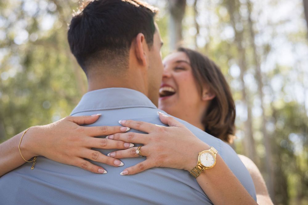 Close-up of a woman laughing while hugging a man outdoors, showing her engagement ring and gold watch.