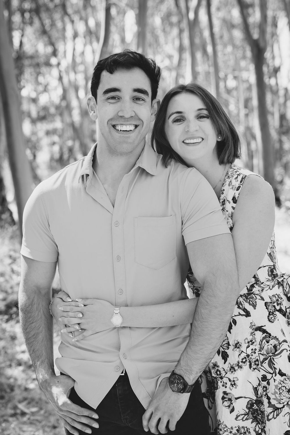 Smiling couple posing together in black and white portrait among tall trees.