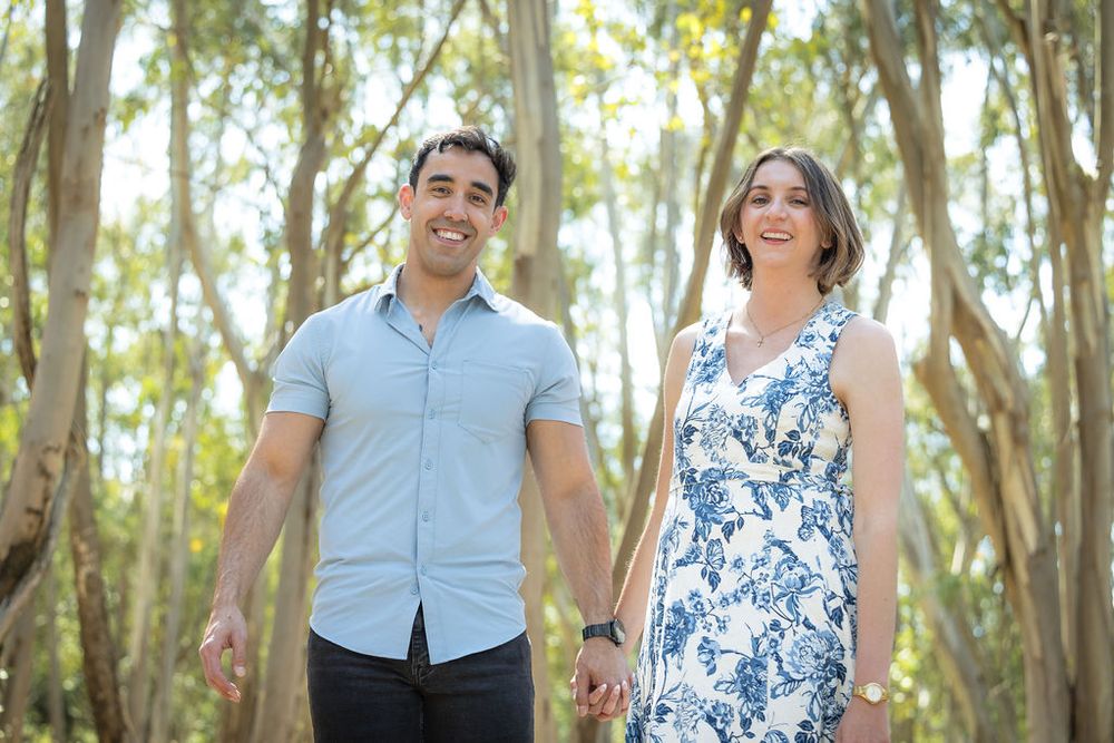 Smiling couple holding hands walking through a eucalyptus forest on a sunny day.