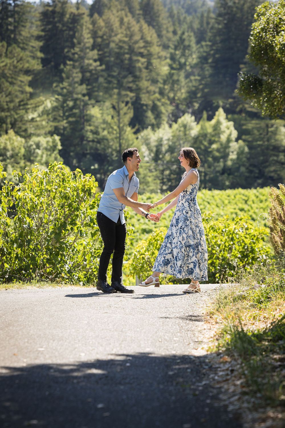 Couple holding hands and dancing on a sunlit road surrounded by lush green vineyard and forest.