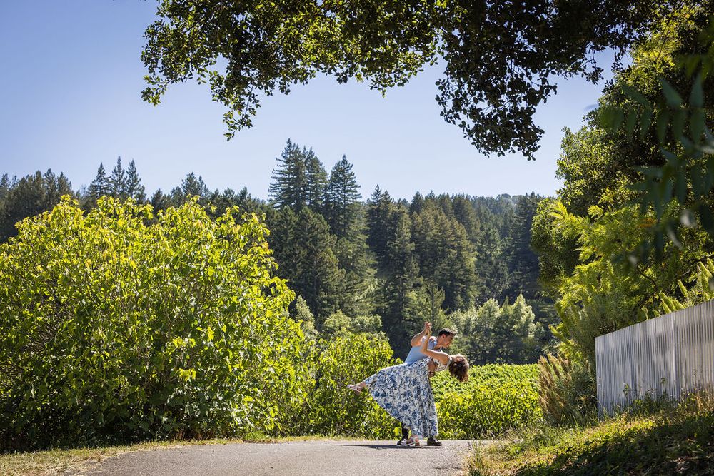 Couple embracing on a sunlit path surrounded by lush greenery and towering redwood trees in a scenic outdoor setting.