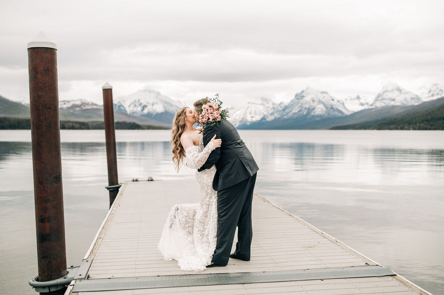 Couple sharing a romantic kiss on a dock at Lake McDonald, Glacier National Park, with snow-capped mountains in the background.