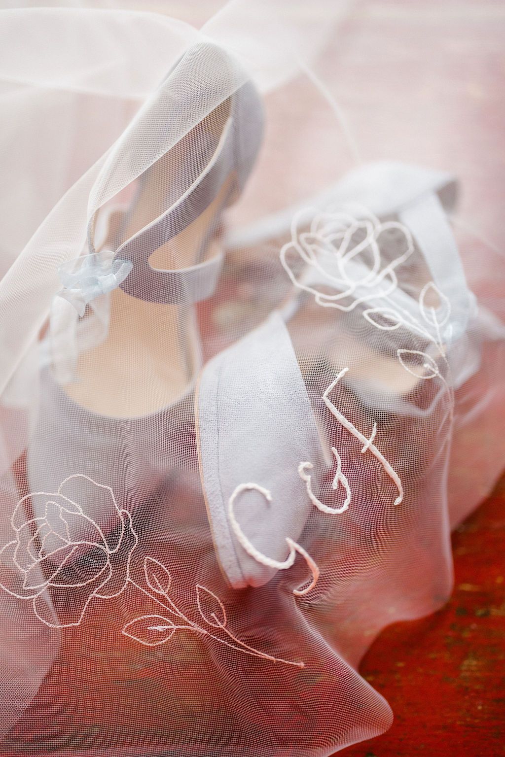 White bridal shoes with monogram embroidery and floral details draped with sheer wedding veil on red surface.