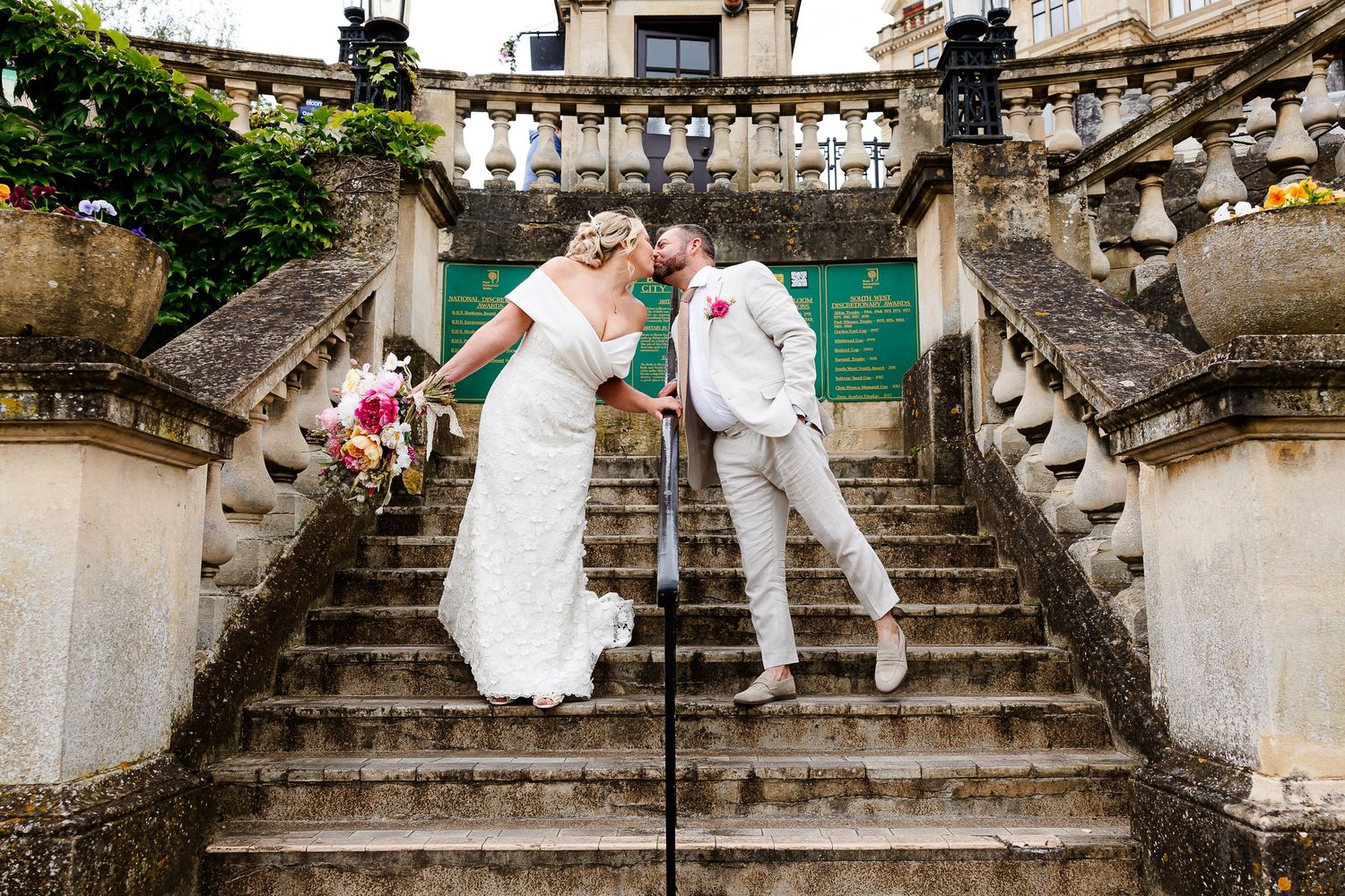 Bride and groom share a romantic kiss on elegant stone staircase at a grand venue on their wedding day.