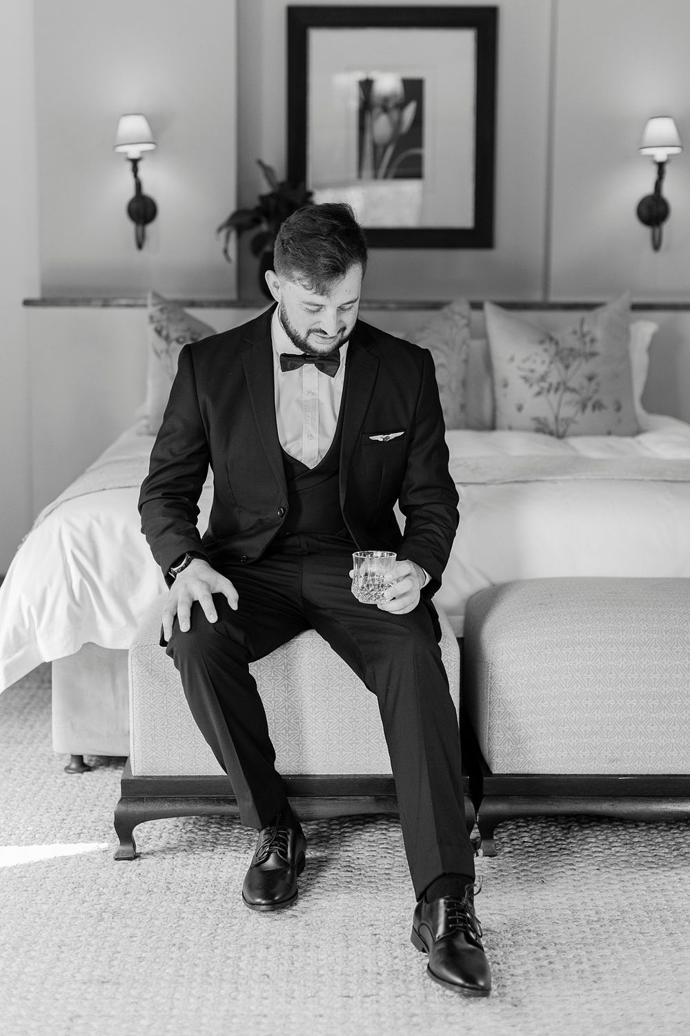 Dapper groom in black tuxedo and bow tie sitting on hotel bed holding a whiskey glass.