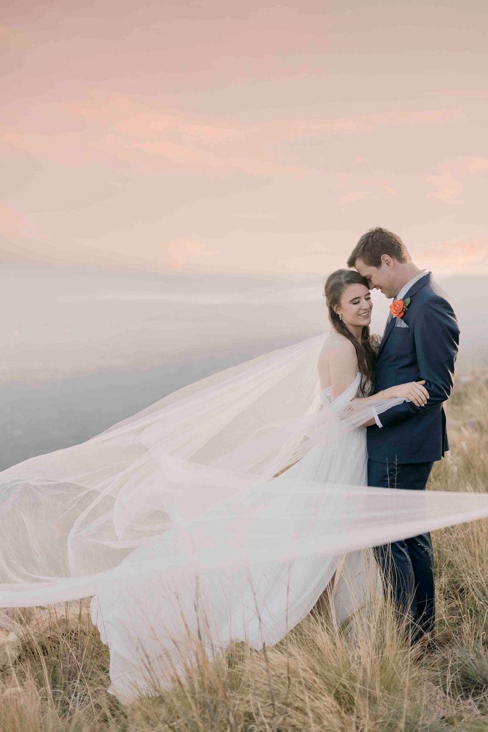 Romantic bride and groom embracing on hilltop at sunset with flowing wedding veil in the wind.