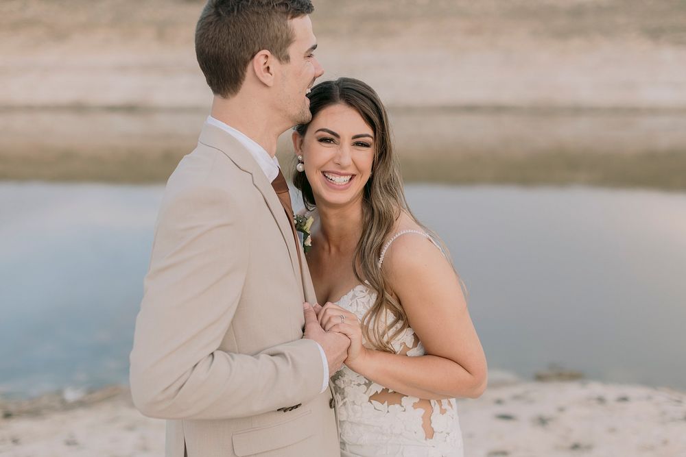 Smiling bride in lace wedding dress embracing groom in beige suit near a serene waterfront.