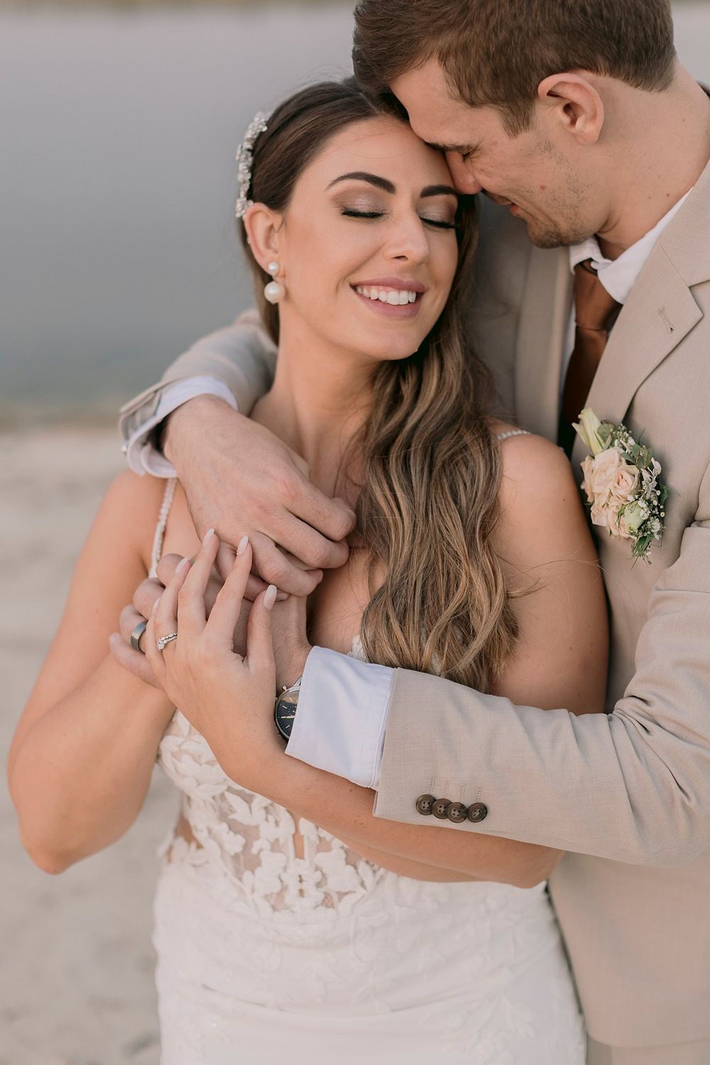 Smiling bride with eyes closed as groom in beige suit embraces her near water.
