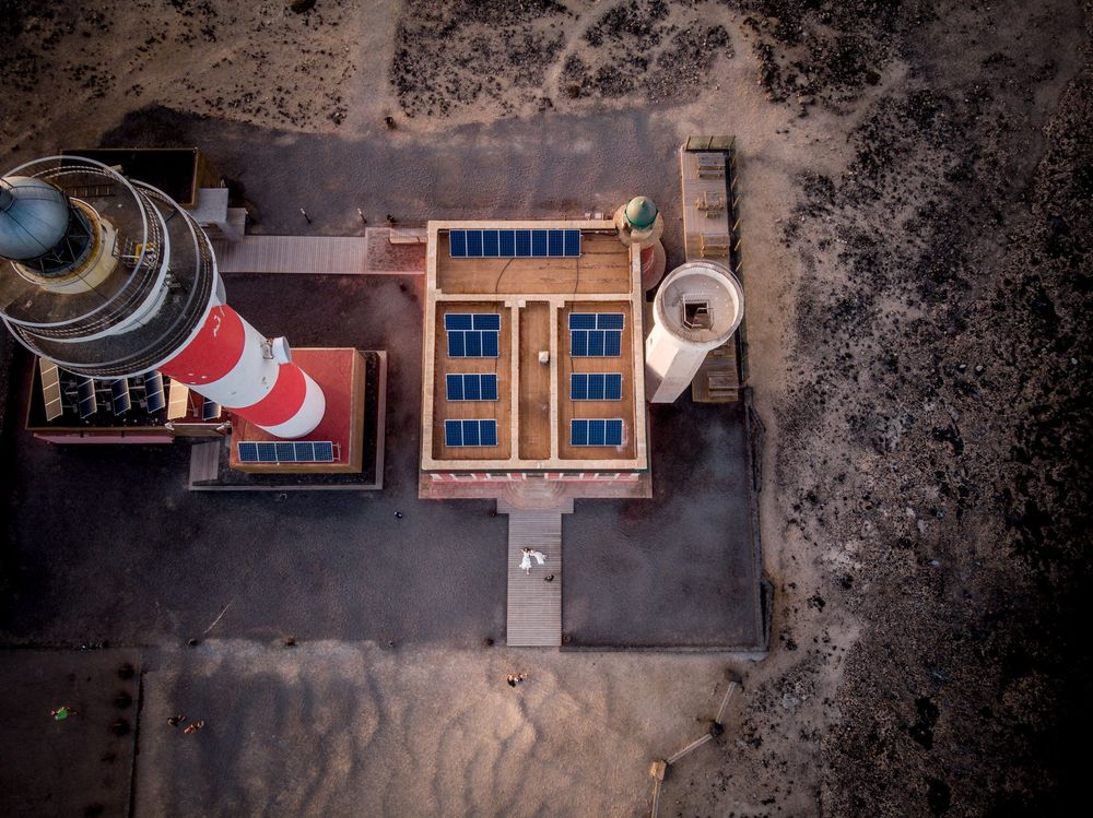 Aerial drone view of a lighthouse with red and white stripes beside a building with blue solar panels.