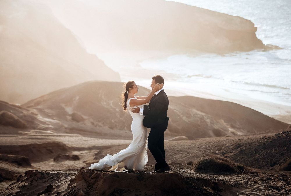 Bride and groom embrace on rocky coastal cliffs with golden sunlight and ocean waves in the background.