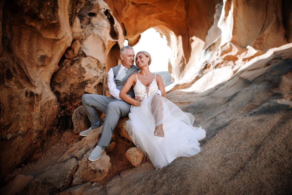 A bride and groom sit together in a stunning red rock canyon, wearing wedding attire in a romantic desert elopement setting.