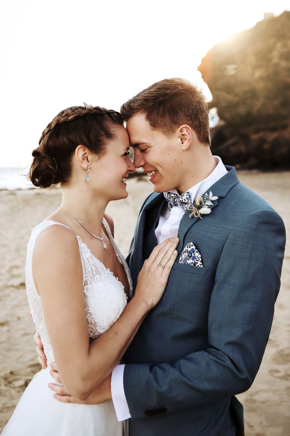 A bride and groom share a romantic forehead touch on a beach, smiling in wedding attire with golden sunset light behind them.
