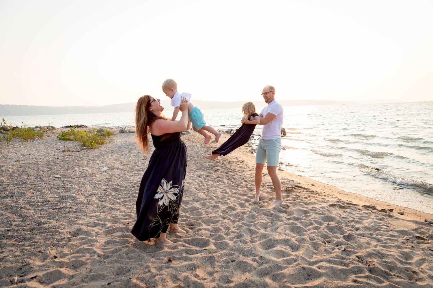 Family of four sitting on rocky beach by water, parents with two young children enjoying sunny summer day outdoors.