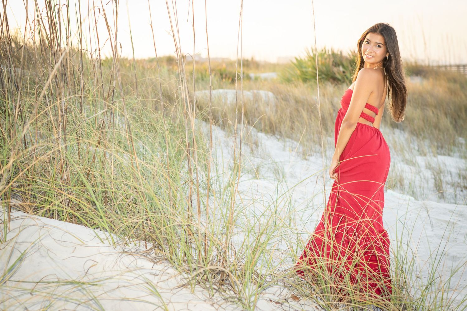 A woman in a red gown poses gracefully on white sand dunes with sea grass at a beach during golden hour.