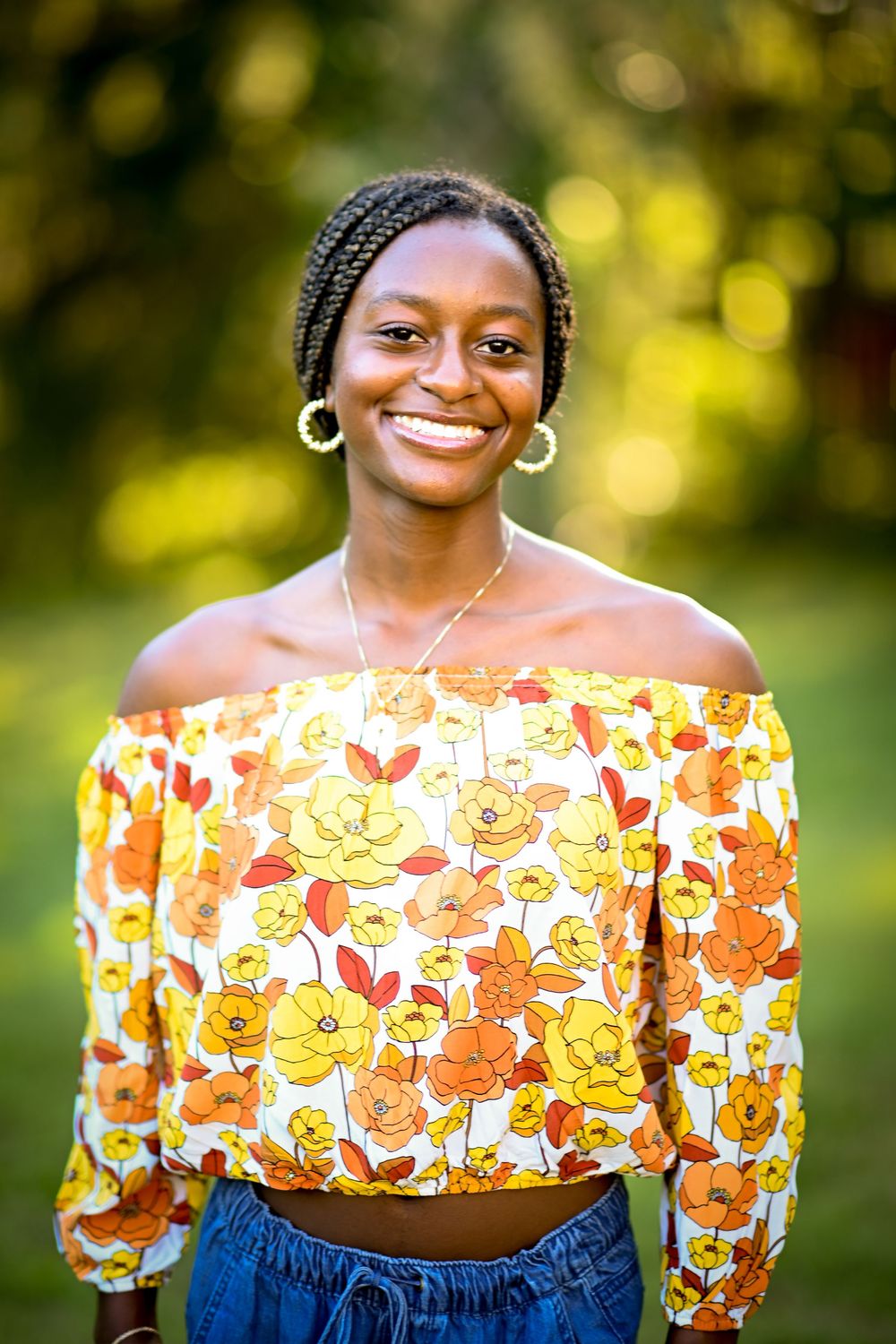Young woman smiling outdoors in floral off-shoulder crop top, braided hair, gold earrings, green bokeh background.