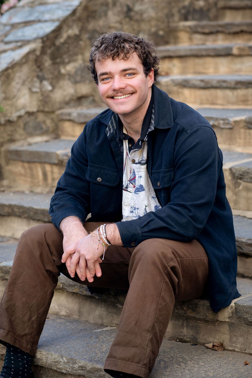 A smiling man with curly hair and mustache sits on stone steps wearing a navy jacket and brown pants.