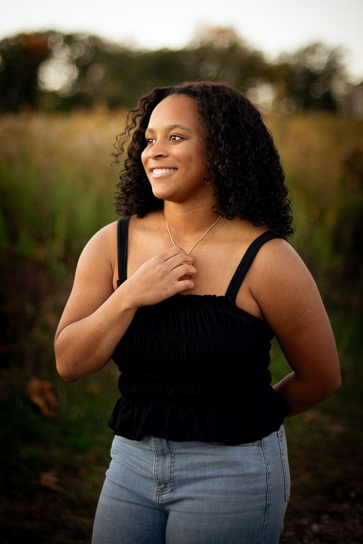 Young woman with curly hair smiling outdoors in a black top and jeans, holding a gold necklace in a sunlit field.