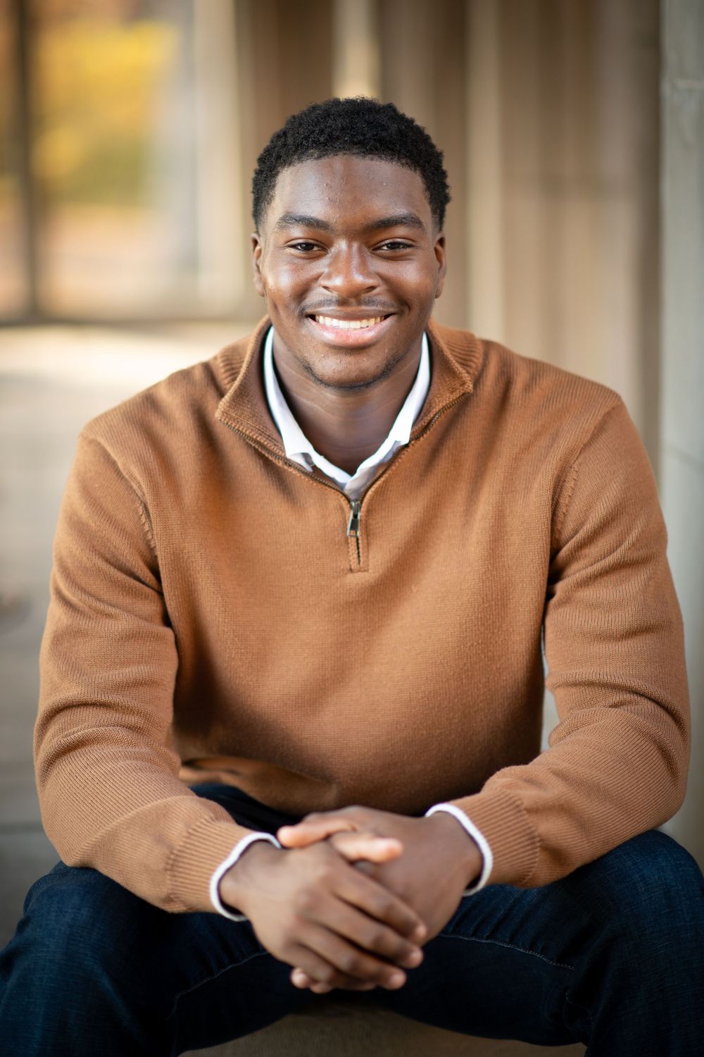 Smiling young Black man in brown quarter-zip sweater sits with clasped hands in an outdoor portrait photo.
