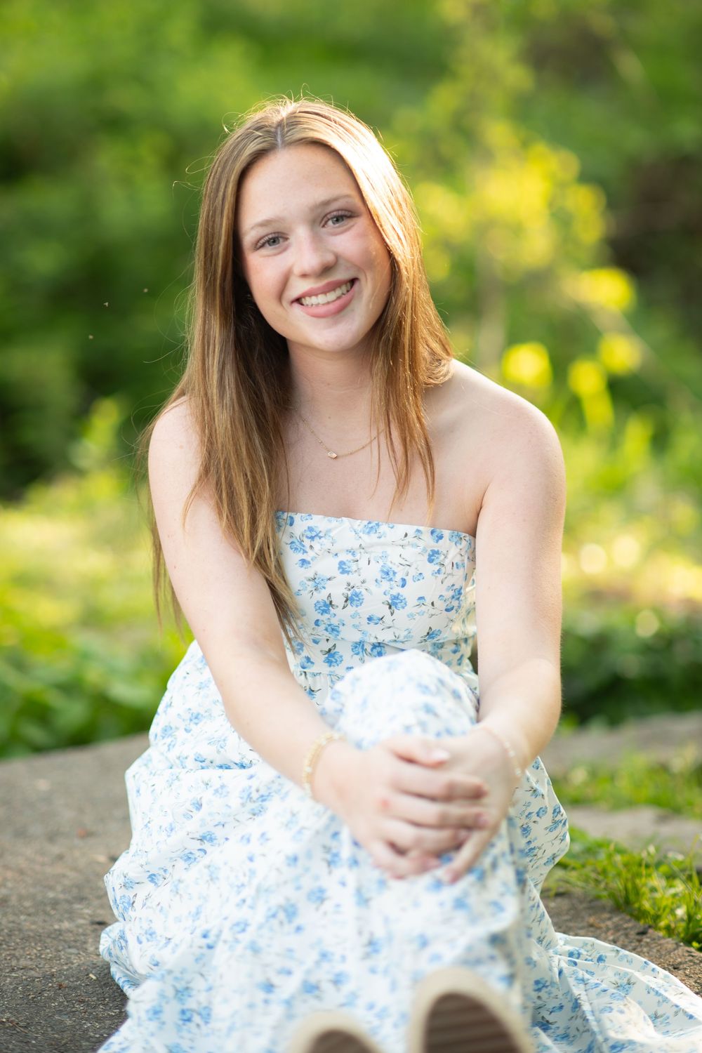 Smiling teenage girl in floral strapless dress sitting outdoors in lush green garden setting.