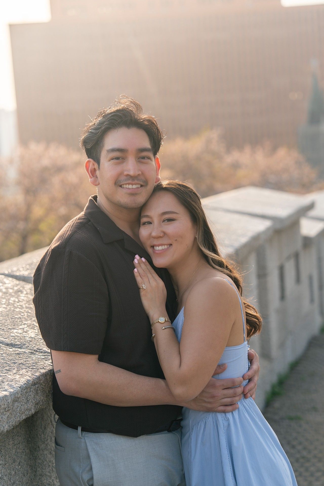 A smiling couple embraces at golden hour on a rooftop, woman in blue dress showing engagement ring.